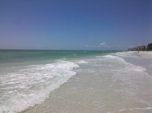 A beach with clear water, a sandy shore, and buildings in the background.