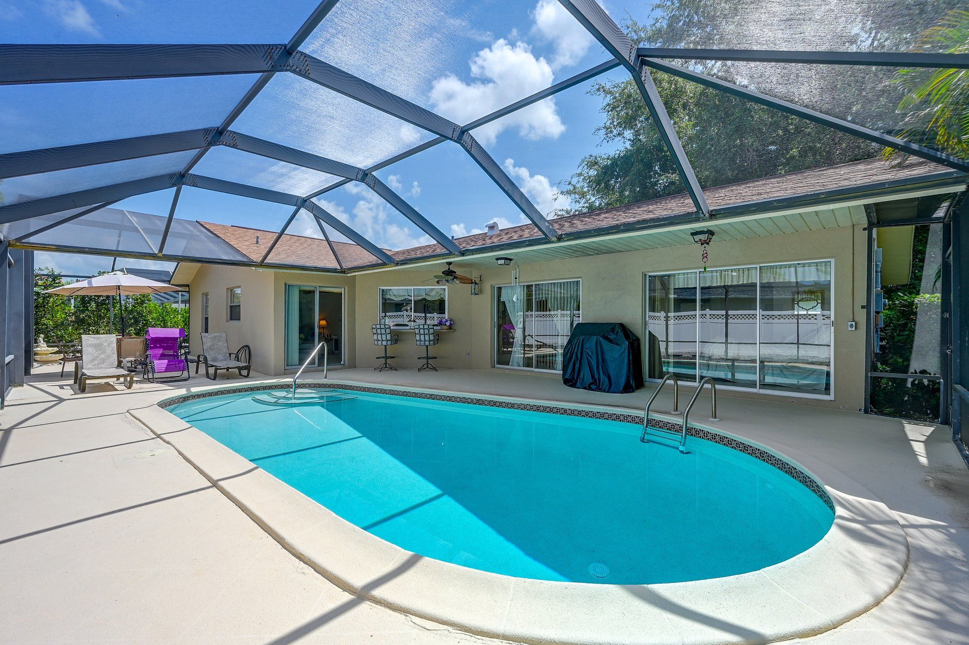 Une piscine entourée de parois en verre, un espace barbecue couvert et une terrasse avec des chaises et des parasols.
