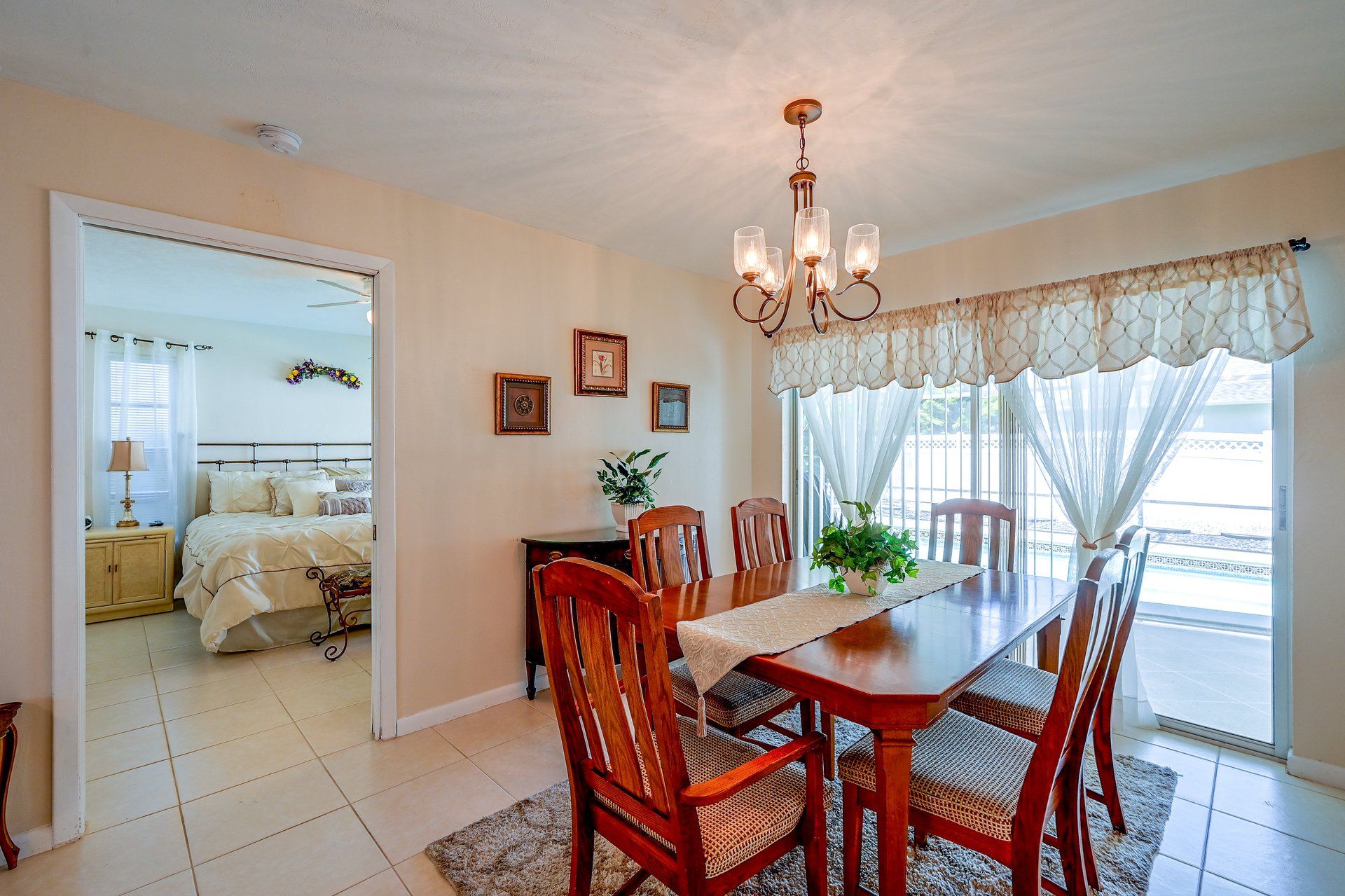 A dining area with a wooden table and chairs, a chandelier, and a view through a window with curtains.