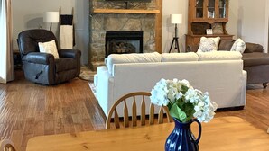 Un salon avec une cheminée en pierre, un téléviseur à écran plat, un canapé, un fauteuil et une table à manger agrémentée d’un vase de fleurs.