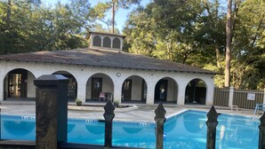 Una piscina con una valla en primer plano, un edificio con arcos al fondo y árboles que rodean el área.