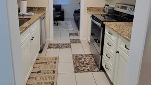 Una cocina con armarios blancos, microondas, estufa y refrigerador. Hay una ventana con persianas y una puerta que da a una sala de estar.