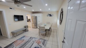 Cocina compacta con armarios blancos, horno empotrado y refrigerador. Hay una mesa de comedor con sillas y un banco. Un televisor está montado en la pared, y se observa un ventilador de techo.