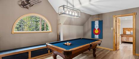 A pool table in a room with a sloped ceiling, a large window with a view of trees, and a wall-mounted light fixture.
