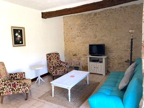 Un salon avec un mur en pierre, un canapé, un téléviseur posé sur un meuble, un fauteuil à motifs et une petite table.