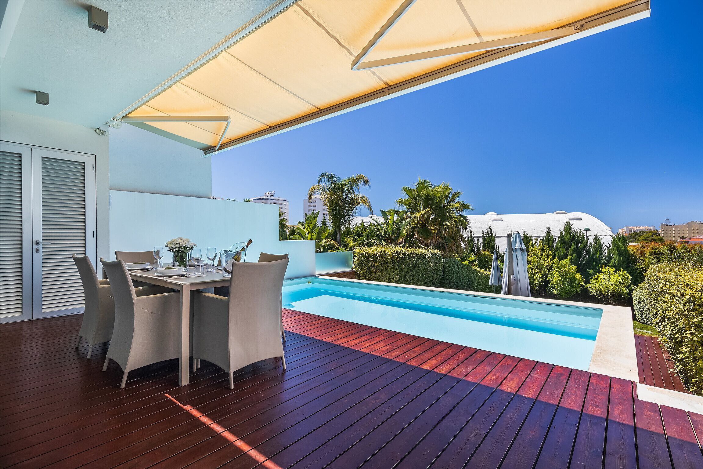 A poolside area with a dining table and chairs, a pool, and a view of palm trees and buildings.