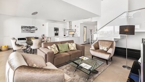 A modern living room with a sofa, armchairs, and a coffee table. There is a dining area with a table and chairs, and a kitchen in the background.