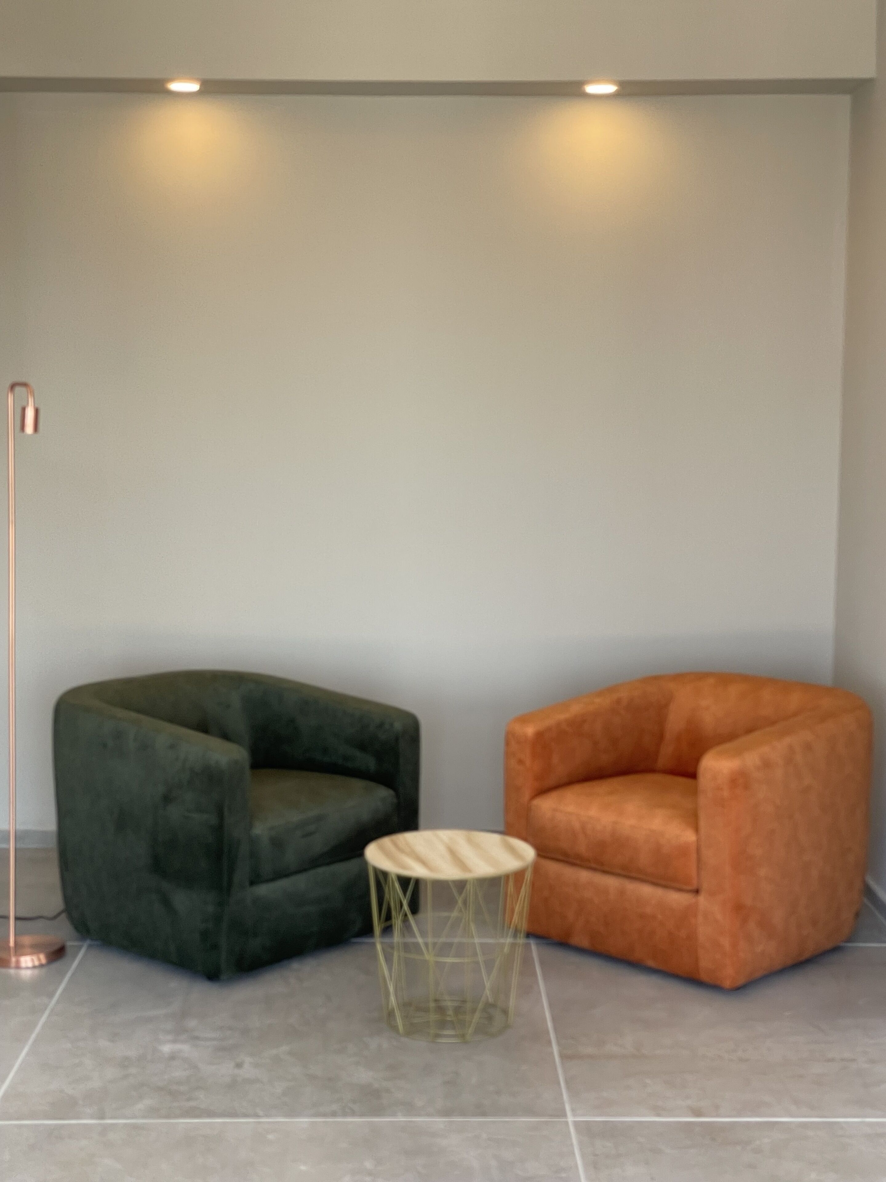 Una habitación con dos sillas, una verde oscuro y otra naranja, y una pequeña mesa redonda con estructura metálica.