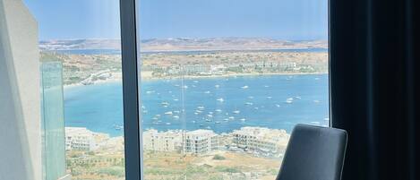 A modern room with a large sliding glass door leading to a balcony, offering a view of a coastal area with buildings and boats.