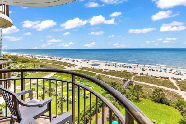 A balcony with a view of a beach, including a pool area and umbrellas.