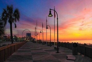 Une promenade en bord de mer, avec des lampadaires et une grande roue en arrière-plan, au coucher du soleil.
