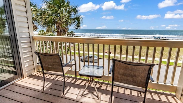 Un balcon avec deux chaises et une petite table, donnant sur une plage bordée de palmiers et sur l’océan.