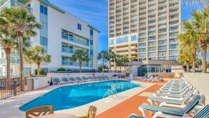 A pool area with lounge chairs and a table, surrounded by palm trees and hotel buildings.