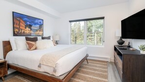 A modern bedroom with a large bed, a window with a view of trees, a painting of a cityscape, and a dark wooden dresser. - Cozy Mountain Village Hotel Room with Jetted Tub (Mountain Village)