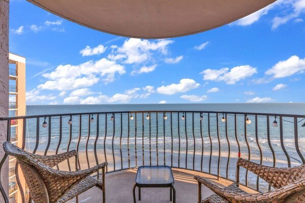Un balcón con sillas de mimbre y una mesa, con vista a una playa con olas y un cielo despejado.