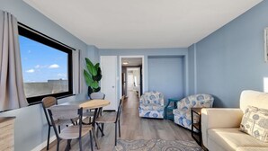 Un salon moderne comprenant un coin repas, un canapé et une table basse. On y trouve une grande fenêtre avec des rideaux, une plante en pot et un tapis à motifs.