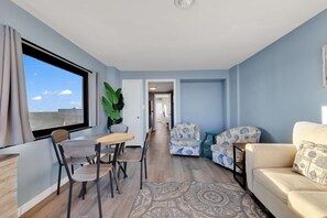 A modern living room with a dining area, a sofa, and a coffee table. There is a large window with curtains, a potted plant, and a patterned rug.