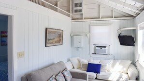 A cozy room with a white wooden ceiling, a white sofa with colorful cushions, a framed picture on the wall, and a window with blinds. - 1 bedroom cottage with ac, steps to the beach! (Dennis Port)