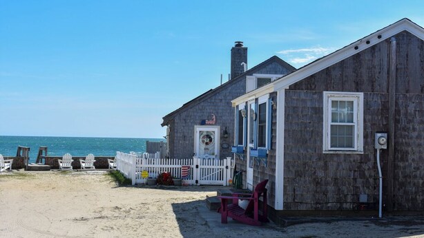 A wooden building with a porch and a white picket fence, located on a sandy beach with the ocean in the background. - 1 bedroom cottage with ac, steps to the beach! (Dennis Port)