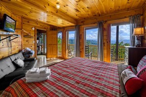 A wooden cabin bedroom with a bed, a sofa, a television, and a balcony with a view. - Mountain-View Cabin w/ Game Loft, Hot Tub & Fireplaces (Sevierville)