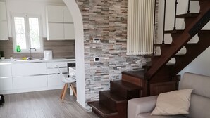 A modern living room with a staircase, a kitchen in the background, and a stone wall. - Nemo's Home (Arcola)