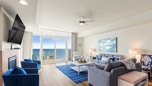 Una sala de estar moderna con televisor de pantalla plana, ventilador de techo, ventana grande con vista al mar y balcón con mobiliario exterior.