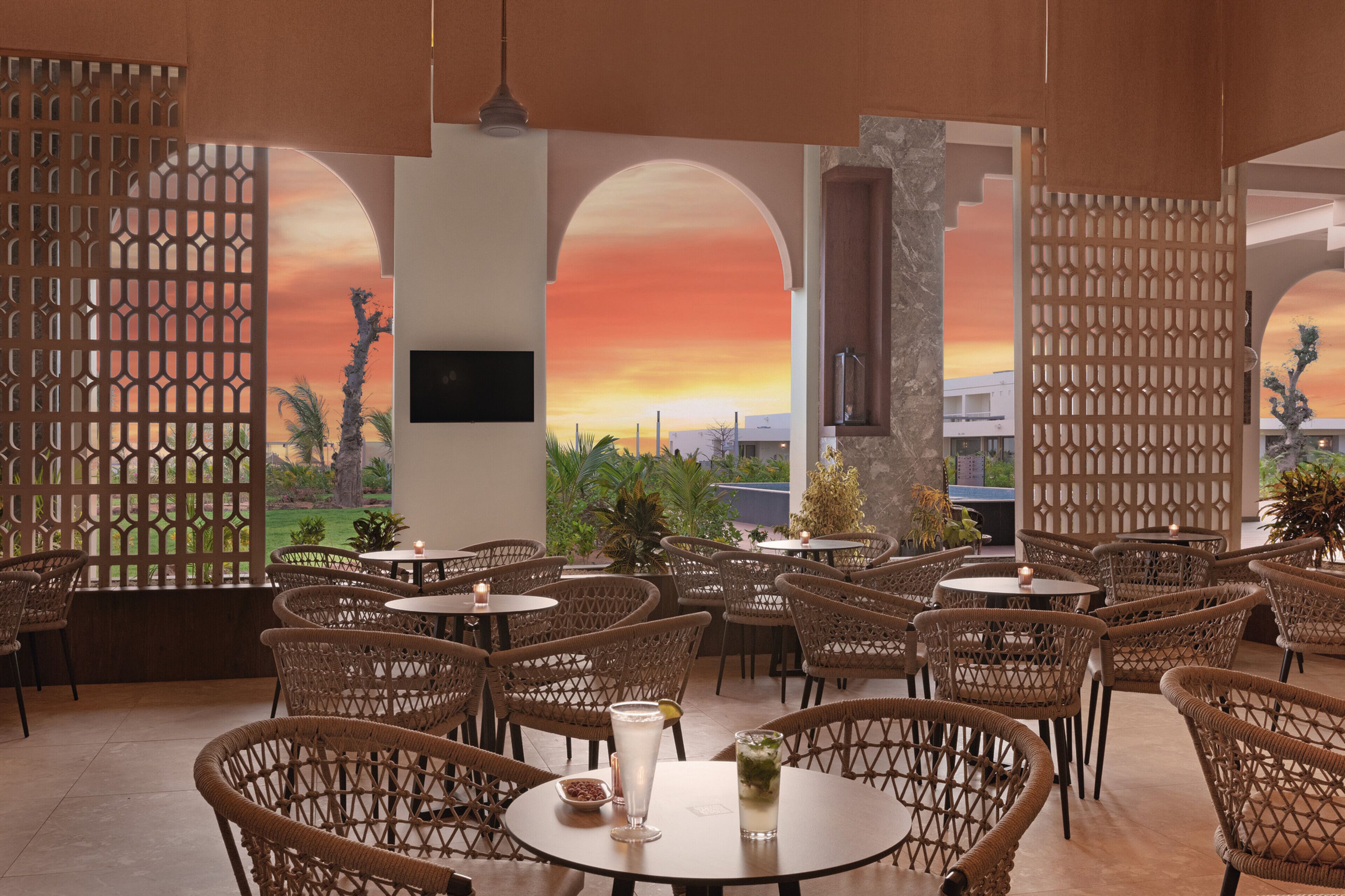 Un restaurante con sillas y mesas de mimbre, vista al atardecer a través de aberturas arqueadas y un biombo decorativo.