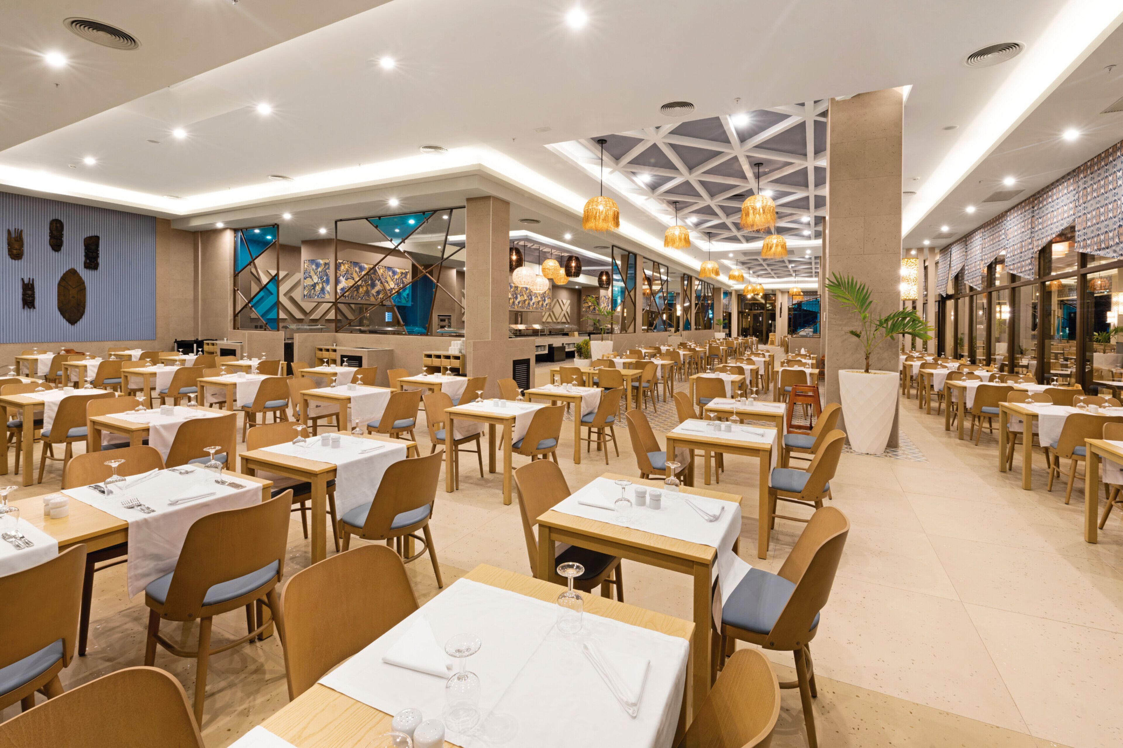 A spacious restaurant with multiple tables and chairs, a high ceiling with geometric patterns, and a modern interior design.