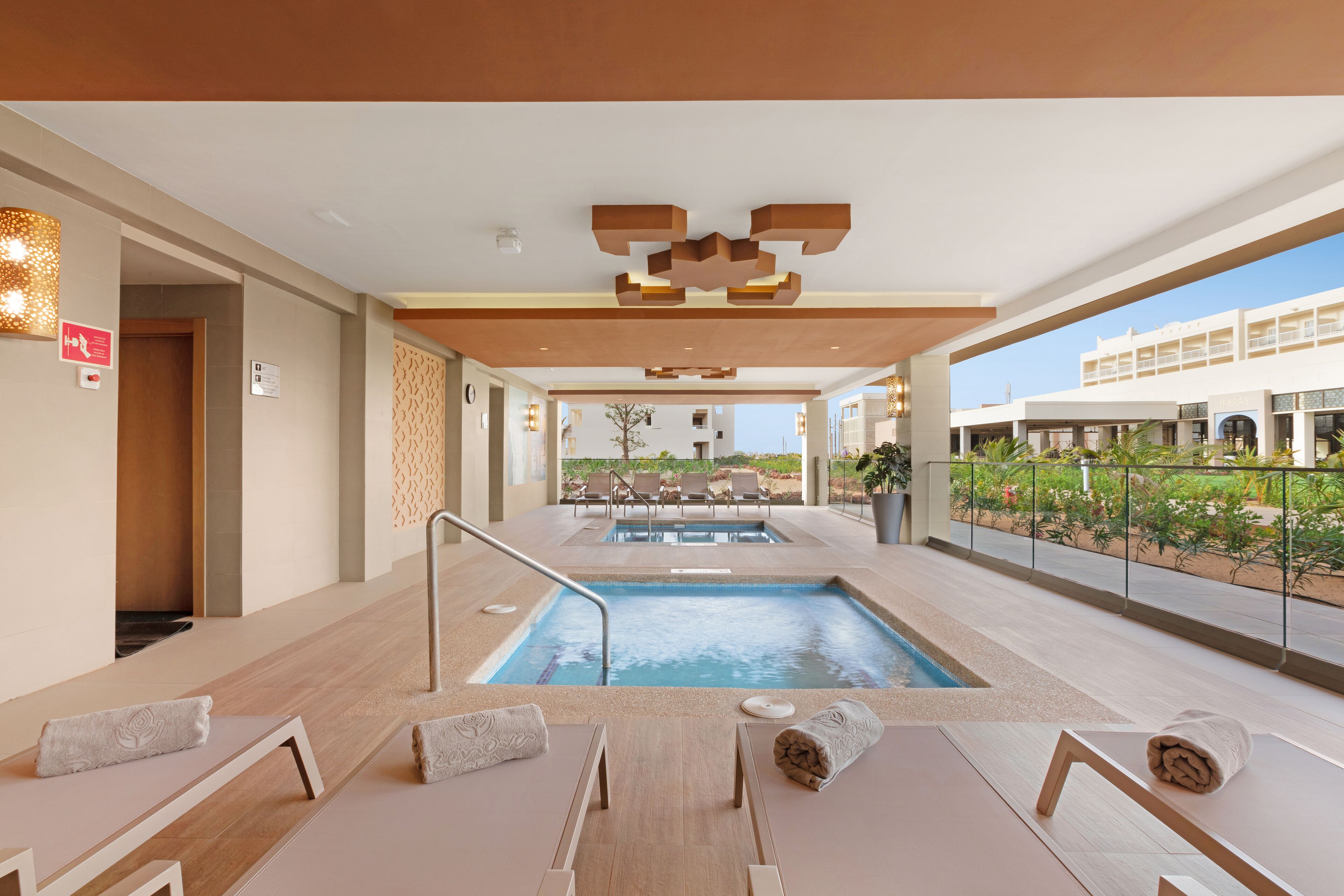 A modern indoor pool area with lounge chairs and a view of the building and greenery outside.