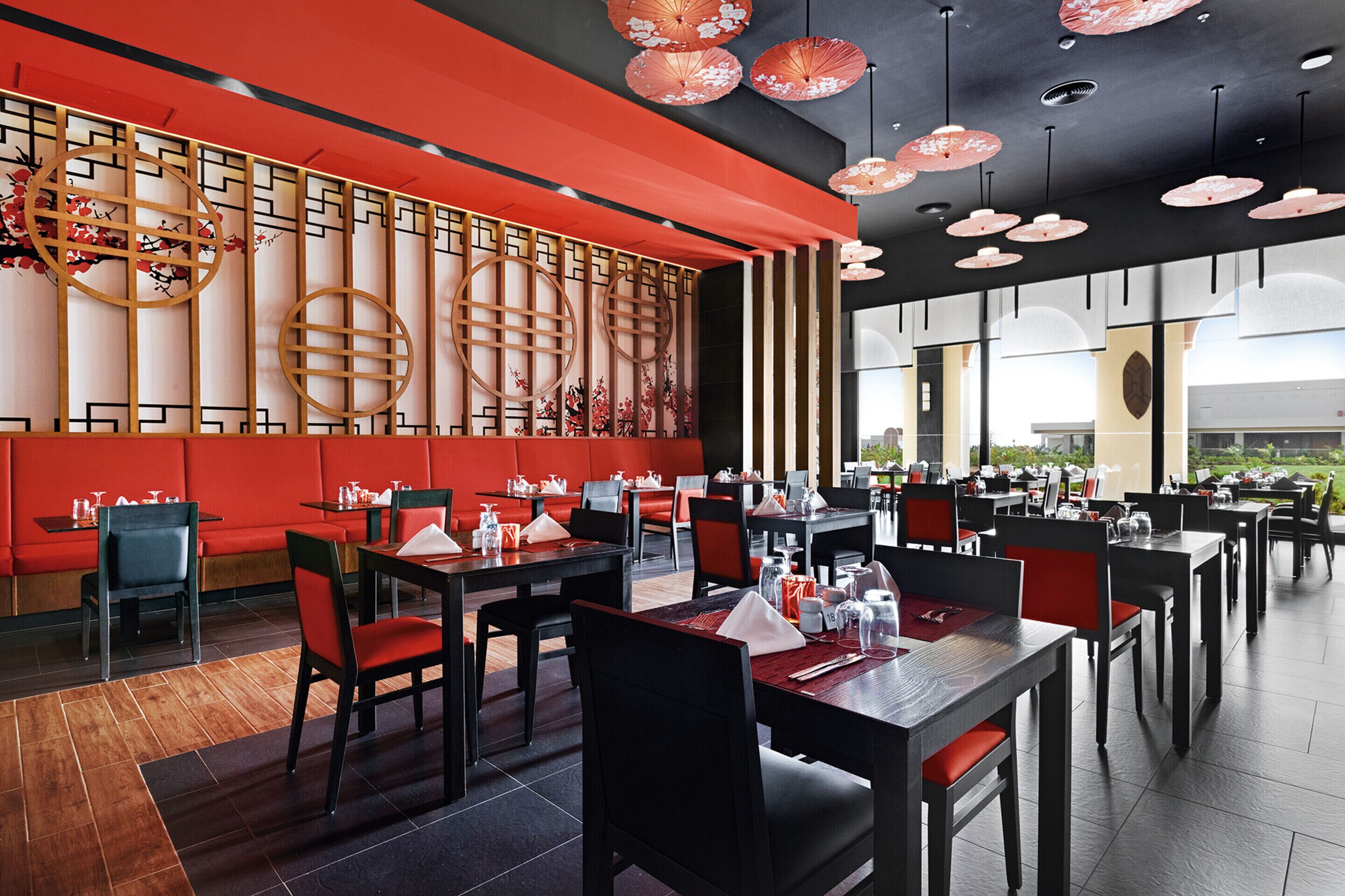 A restaurant with red seating, wooden decor, and hanging red umbrellas. Tables are set with white tablecloths and red chairs.