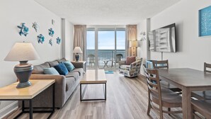 A modern living room with a sofa, coffee table, and dining table, a balcony with a view, and decorative wall art. - Avista Resort 823-1BR/1BA Oceanfront Condo in Ocean Drive (North Myrtle Beach)