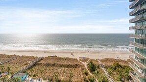 Vista de una playa con un paseo marítimo, una torre de salvavidas y un edificio alto con balcones.