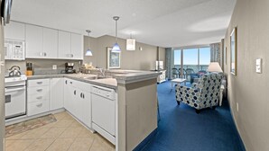 A modern kitchen with white cabinets, a dishwasher, and a large island. There is a dining area with a patterned chair and a view of the ocean through large windows.