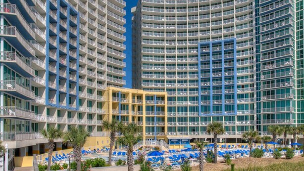 A multi-story hotel with balconies, a pool area with sun loungers, and palm trees.
