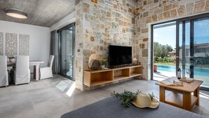 A modern living room with a stone wall, a flat-screen TV, a dining area with chairs, and a view of a swimming pool. - Villa Angelos - Three Bedroom Villa, Sleeps 6 (Ionian Islands)