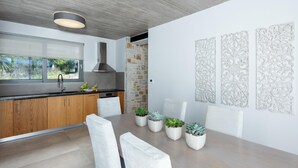 A modern kitchen with a large island, wooden cabinets, and a stone accent wall. There are three potted plants on the island and a dining table with chairs. - Villa Angelos - Three Bedroom Villa, Sleeps 6 (Ionian Islands)