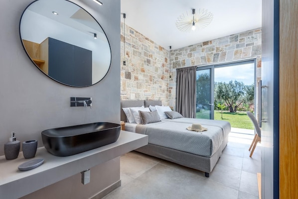 Une chambre moderne avec un grand lit, un miroir rond, un lavabo noir et un mur en pierre.