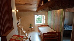 Une pièce au plancher de bois, avec un lit simple, une petite table et une chaise, une fenêtre et un escalier.
