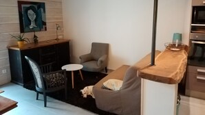 Un salon moderne avec un canapé, un fauteuil, une petite table ronde et un chien. Il y a un tableau au mur et un coin cuisine avec un four à micro-ondes et un évier.