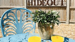 A blue metal chair, a potted plant, and two glasses of orange juice on a table with a wooden fence in the background. - The Hideout (Chichester)