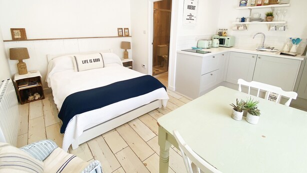 A compact bedroom with a bed, bedside tables, a nightstand, a small seating area, and a kitchenette with a dining table and chairs. - The Hideout (Chichester)