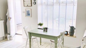 A dining area with a green table and white chairs, a window with blinds, and a small shelf with plants and a kettle. - The Hideout (Chichester)