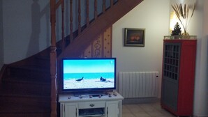 A living room with a wooden staircase, a television displaying a beach scene, a white cabinet with figurines, a red cabinet, and a dining table with a chair. - House 6 pers. direct beach access(100 meters walk) (Borgo)