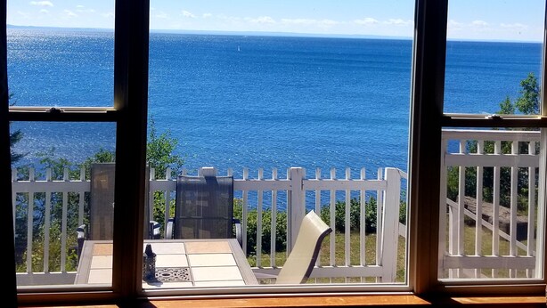 A view of the ocean through a window with a white railing and a table with chairs.