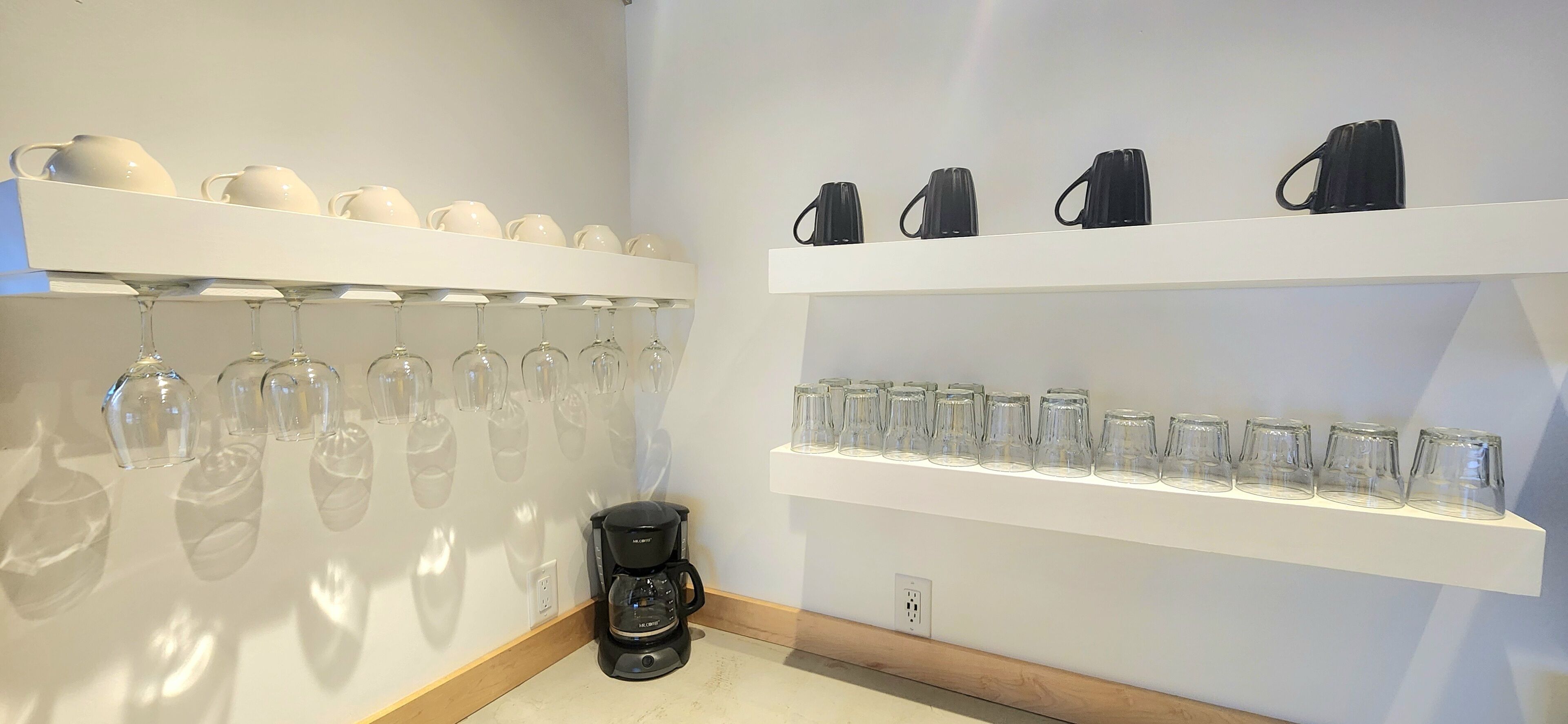 Een kamer met twee witte planken waarop divers glazen servies en een koffiezetapparaat op de vloer staat.