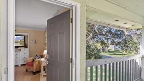 An open door leading to a room with a desk and chair, a window with a view of a garden, and a balcony with a railing.