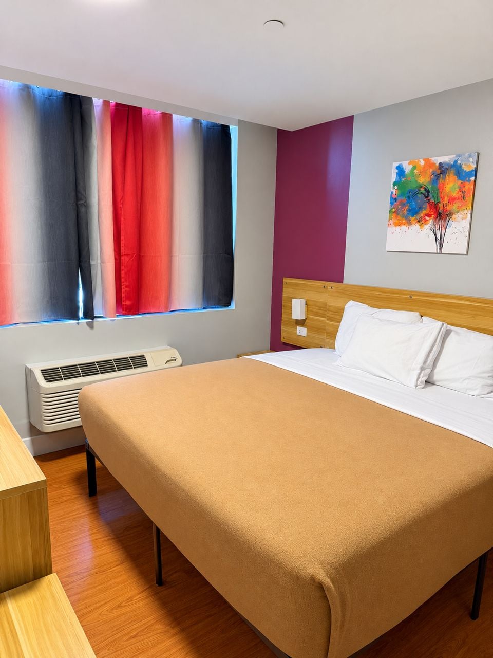 A hotel room with a bed, a bedside table, a window with curtains, an air conditioner, and a wall-mounted painting.