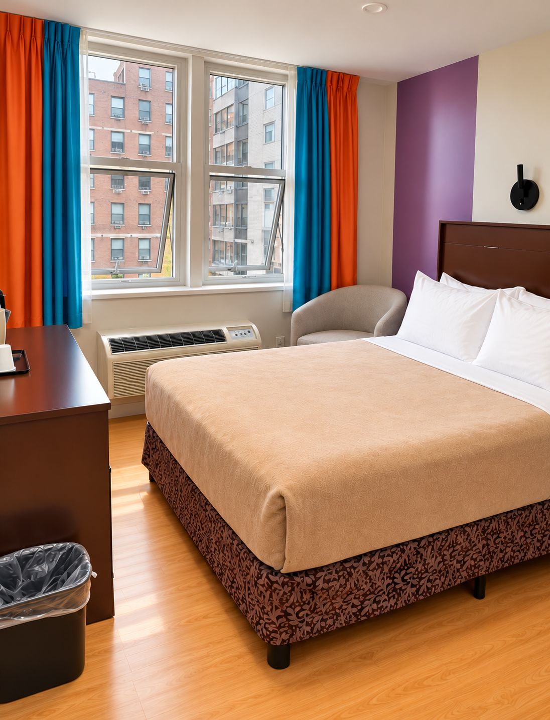 A hotel room with a bed, a desk, an air conditioning unit, a chair, and a view of buildings through the window.