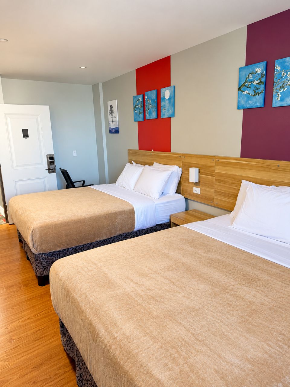 A hotel room with two beds, a wooden headboard, a white door, and artwork on the wall.