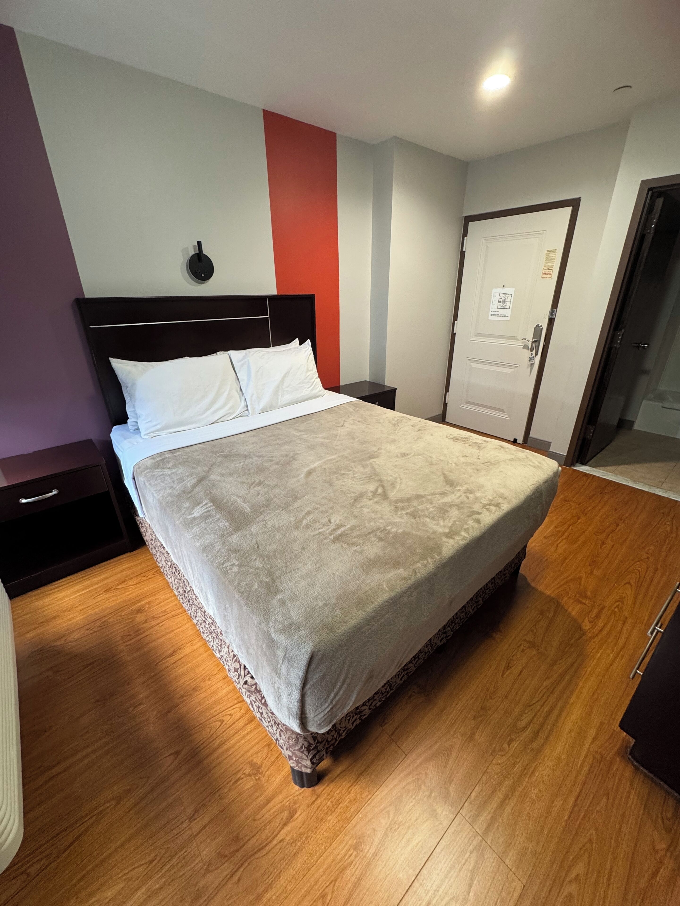 Una habitación de hotel con una cama grande, una mesita de noche, una puerta y un aplique de pared.